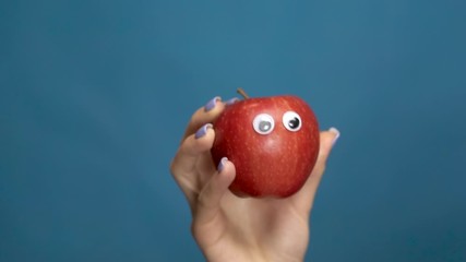 Red apple with eyes in a woman hand close-up. Apple shakes and twists eyes on a blue background. Slow motion.