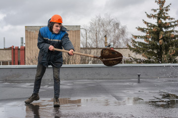 Worker builder on the roof of the factory removes water from the puddle with a shovel. on the head a construction helmet. work at height.