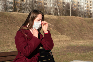 girlis sitting on a bench in the street. puts on a face a white protective medical mask. protection against viruses, coronavirus transmitted by airborne droplets. dressed in a red coat