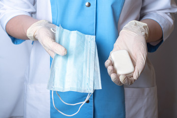 The nurse is holding a white scented soap and protective face masks. Personal hygiene items. Hands are wearing disposable medical gloves. Remedies for viruses, diseases, flu and coronavirus