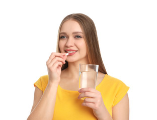 Young woman with glass of water taking vitamin pill on white background