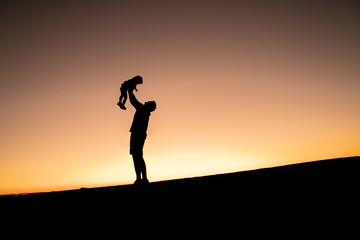Father's day. Silhouette of a happy joyful father having fun throws up in the air little cute child baby girl on nature, sunset horizon background.