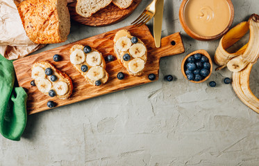 Buckwheat healthy bread with peanut butter, banana and blueberry on wooden board over concrete background. Top view. Flat lay. Summer breakfast.
