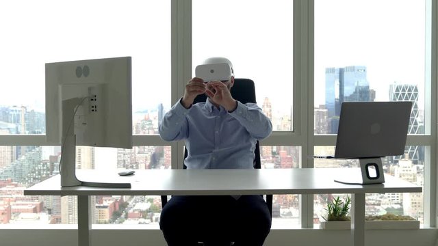 Caucasian male employee sitting at desktop in futuristic office and doing haptic toughing using virtual reality headset, concept of work with 3D dimension for creating digital VR blueprints