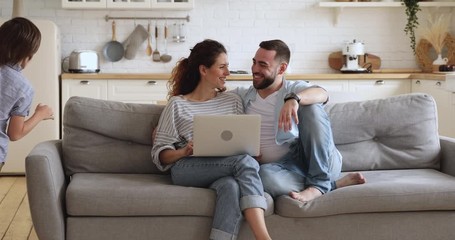 Happy family or four enjoy weekend lifestyle relaxing in modern kitchen interior, young parents couple sit on couch using laptop funny little cute active kids children running playing in modern home - Powered by Adobe