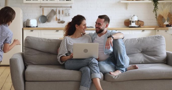 Happy family or four enjoy weekend lifestyle relaxing in modern kitchen interior, young parents couple sit on couch using laptop funny little cute active kids children running playing in modern home