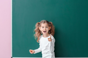 Cute little schoolgirl with raised index finger near blackboard in classroom