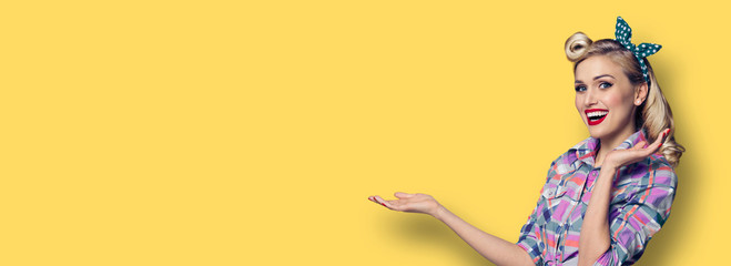 Woman holding, pointing or giving something. Excited adorable girl in pin up style, showing some product or copy space for slogan or text. Retro fashion and vintage, over yellow background.