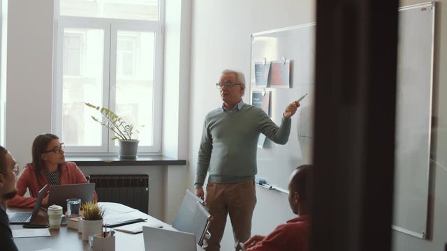 Senior Caucasian male manager pointing at graph on white board and giving presentation to team of young multiethnic employees during corporate training