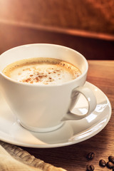 Eine Kaffeetasse mit gerösteten Kaffeebohnen auf einem hölzernenen Tisch. Sonneneinstrahlung, Reflexionen. Ein Cappuccino mit Milchschaum und gemahlenem Kaffee. Serviert in einem Café. Milchkaffee