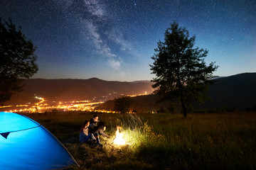 Happy couple hikers resting at campfire near illuminated blue tourist tent under amazing night sky full of stars and Milky way. On the background starry sky, mountains, big tree and luminous town