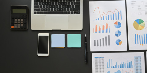 Top view image of Computer laptop, black empty screen smartphone, calculator, post it, graphic charts and paper work flat lay on black working desk. Orderly work space concept.