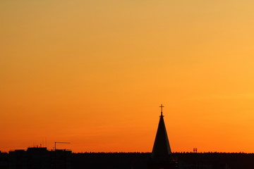 Fototapeta na wymiar Sun below the horizon and the roof of the church with a cross on the background fiery dramatic orange sky at sunset or dawn backlit by the sun. Place for text and design.