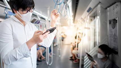 Asian man wearing surgical face mask using smartphone on skytrain or urban train. Wuhan coronavirus (COVID-19) outbreak prevention in public transportation. Health awareness for pandemic protection