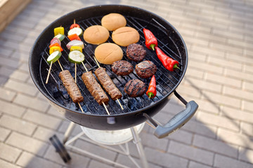 cooking, barbecue and food concept - close up of shish kebab meat, vegetables on bamboo skewers, burger buns and cutlets roasting on brazier grill outdoors