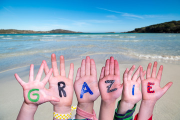Children Hands Building Colorful Italian Word Grazie Means Thank You. Ocean And Beach As Background