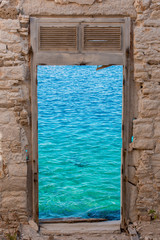 Window with wooden frame, partly destroyed blinds on the top, retro light brown brick wall close up, clear blue sea surface on background