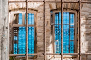 Fototapeta na wymiar Rusty wrought iron fence on foreground, two windows with iron fences deeply blurred, turquoise see water surface with ribbles on background