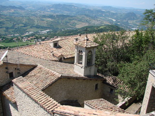 Fototapeta na wymiar View from above of the tiled roofs of ancient houses of a small state under the rays of the summer sun surrounded by garden trees.