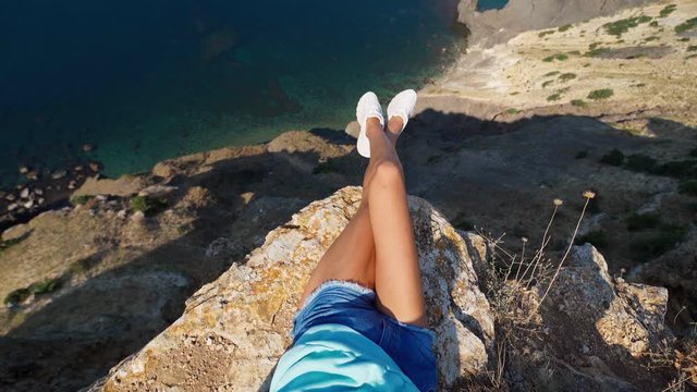 POV first person view taned woman legs in white sneakers and jeans shorts sitting on cliff edge with amazing view seascape with high limestone cliff over blue sea. Traveler enjoying recreation travel