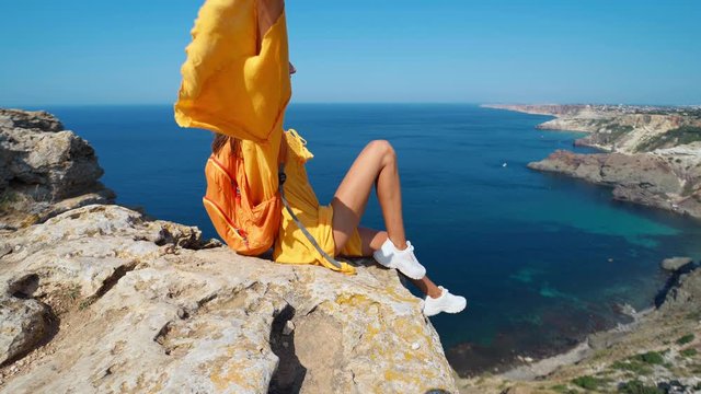 taned traveler woman sitting on top rock beach with raised hands in front of amazing seascape, bright yellow dress and hair blowing in the wind. Freedom, travel and vacation concept.