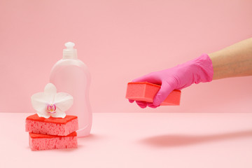 Bottle of dishwashing liquid, sponges and hand with a sponge on a pink background.