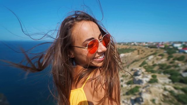 closeup clow motion portrait beautiful young smiling brunette woman with long wind blowing hair. girl wearing in orange sundress and sunglasses, wind flying hair to camera