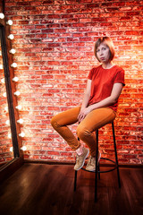 Girl in red shirt on a tall chair in front of a lighted mirror in a dark room. Model posing for a...