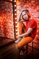 Girl in red shirt on a tall chair in front of a lighted mirror in a dark room. Model posing for a...