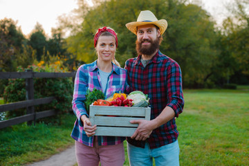 A man and a woman holding a box with a crop of farm vegetables background of the garden.