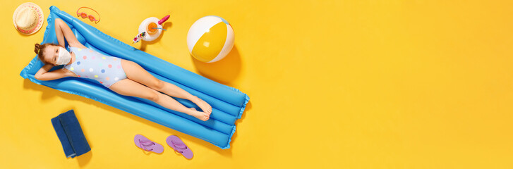 Top view on a little girl in a swimsuit and a protective mask resting on an inflatable blue mattress on a yellow background, concept of summer vacation. Banner copy space