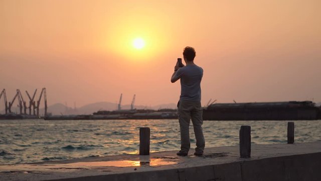 Man Taking Photos of Sunset over Ocean and Harbour With Smartphone