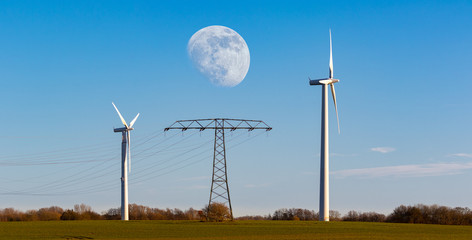 Two wind turbines for wind energy and an electricity pylon with cables in the middle on a field on the Baltic island of R&uuml;gen. It is just before sunset and the moon is in the sky.
