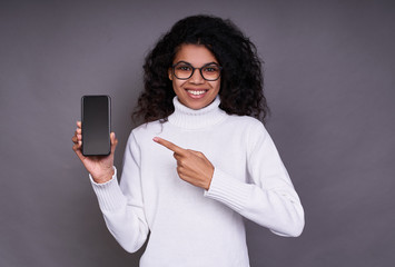 Smiling cute young african american is pointing finger at smartphone in hand.