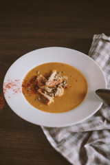 White plate with pumpkin puree soup, sunflower seeds and crackers added to the soup. Topped with chili pepper. Next to the plate is a linen towel of natural colors and a spoonful of silver.