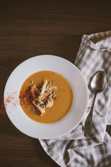 Pumpkin puree soup with crackers and seeds sprinkled with chili peppers in a large plate on a dark wooden background. Nearby lies a silver spoon and a linen towel. Healthy and wholesome food. View fro