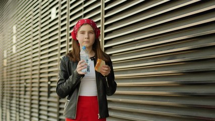 Teenager girl kid walks in a city street eating tasty sandwich drinking water on the go from plastic bottle