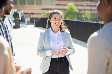 Smiling Caucasian woman talking to friends on street. Cheerful young business consultant talking with clients while standing outdoors. Communication, business concept