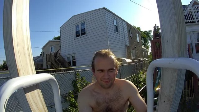 Slightly obese man recording hims self walking and climbing swimming pool leader and jumping into it while bright sun shines behind him.