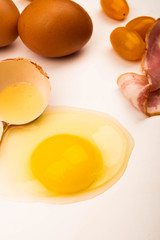 Broken chicken egg, tomatoes, slices of sausage and bacon and chicken eggs scattered on a white background. Close up.