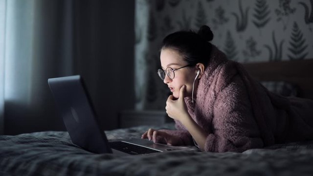 woman lies on the bed home with laptop. girl student wear headphone study online with internet teacher learn language talk looking at laptop, focused young woman make video call
