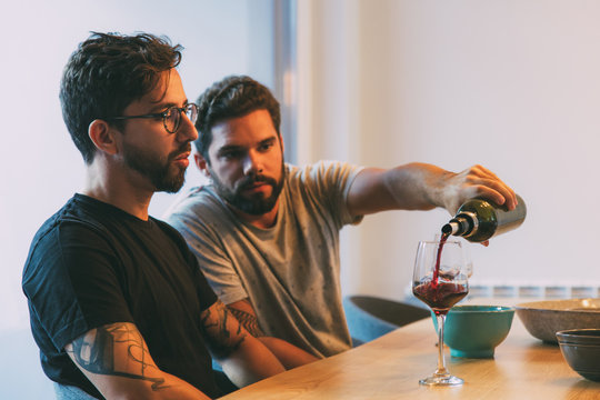 Serious bearded guy pouring wine for his upset friend in dining room. Young men and women in casual meeting indoors. Friendly support concept