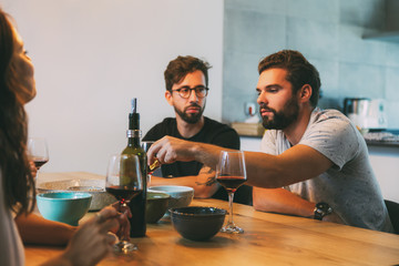 Group of friends having dinner with snacks and wine in dining room. Young men and women in casual meeting indoors. Friendly celebration concept