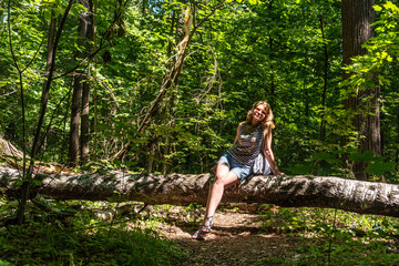 Portrait of an attractive adult woman sitting on a fallen tree on a sunny day against the background of a green forest