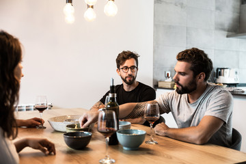 Group of friends chatting at dining table, drinking wine, eating snacks. Young men and women in casual meeting indoors. Friends gathering together concept