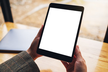 Mockup image of a woman holding black tablet pc with blank white screen with laptop on wooden table