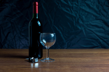 Botella de vino tinto junto a copa de cristal  y tapón de metal en mesa de madera y sobre fondo oscuro.