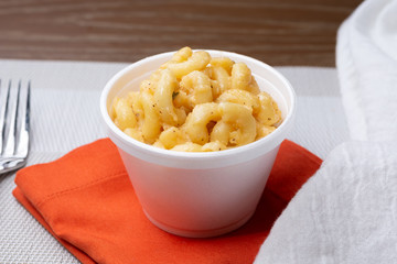 A closeup view of a styrofoam cup of macaroni and cheese, in a restaurant or kitchen setting.