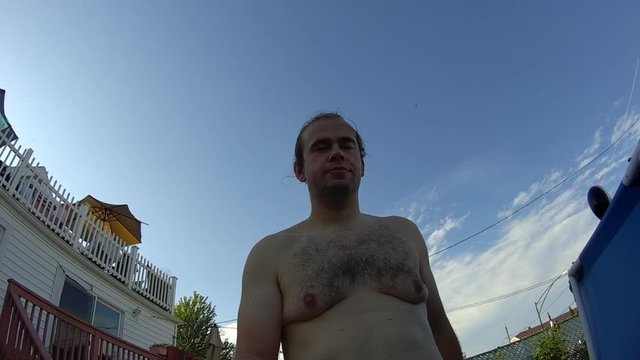 Slightly obese young adult male recording hims self walking and climbing swimming pool leader and jumping into it while bright sun shines behind him.