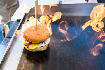 A large white hamburger with bacon and egg is cooked on the stove, grilling with a flame of fire..
